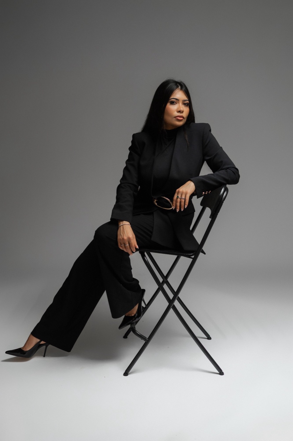 Asenate Pereira, digital marketing strategist, in a black suit seated on a black chair in a grey studio