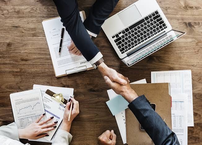 Two business people shaking hands over a desk with a contract and a laptop